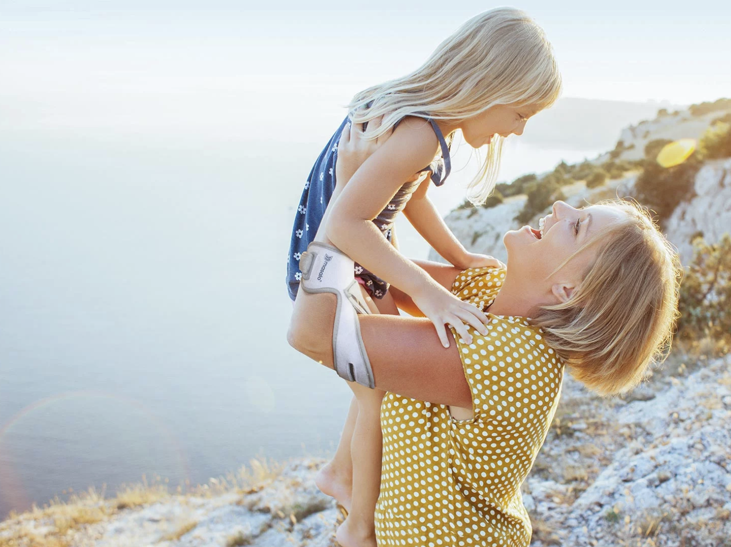 The Masalo Cuff – for more quality of life in everyday life, work & sports Young woman wearing the Masalo cuff is lifting her young daughter into the air. Both are standing on a cliff by the sea.